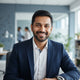 A friendly South Asian man smiling warmly in a professional office setting with soft blue and white tones.